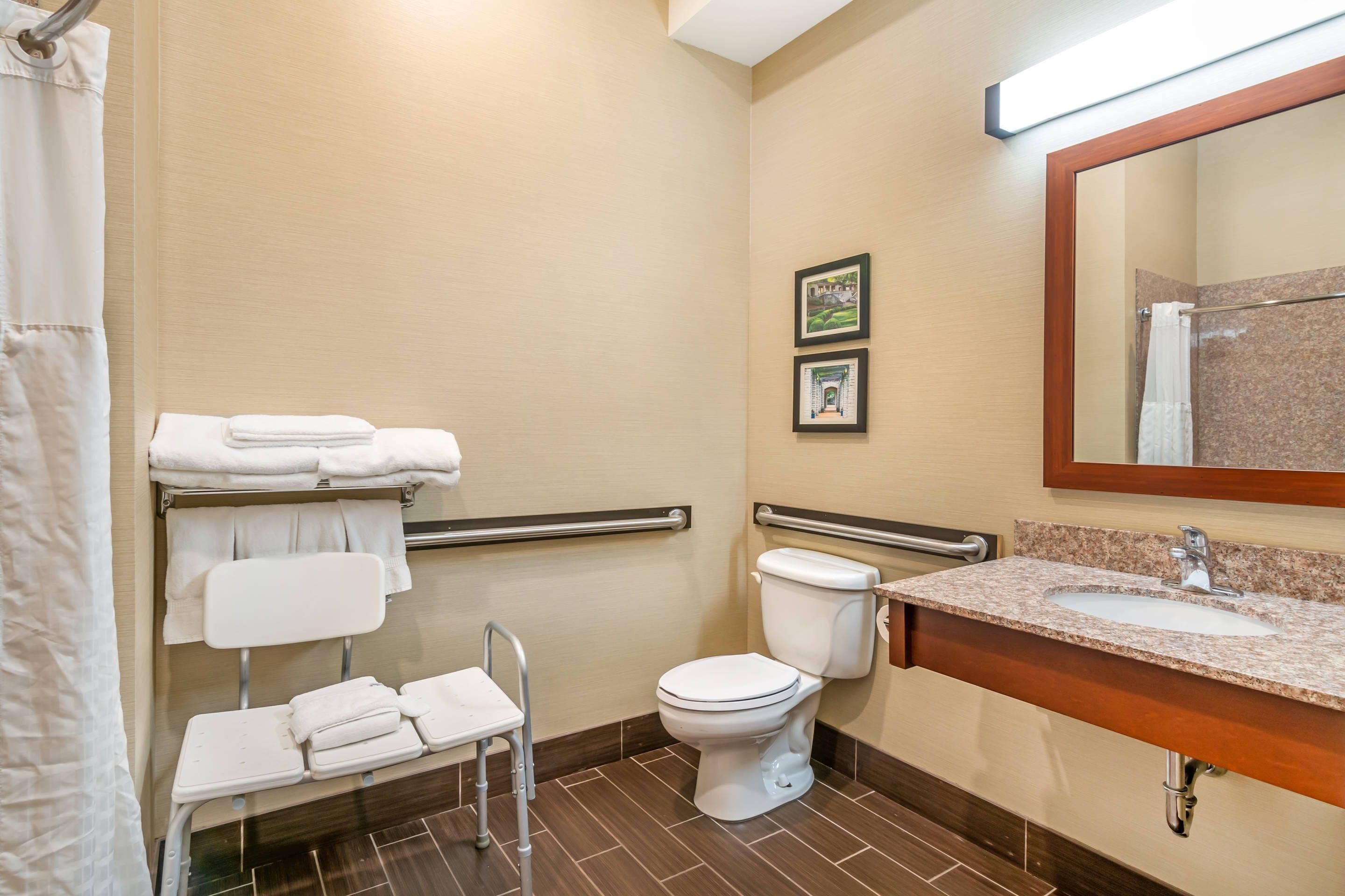 Un baño con inodoro, lavamanos, espejo, cortina de ducha y una silla con toallas.