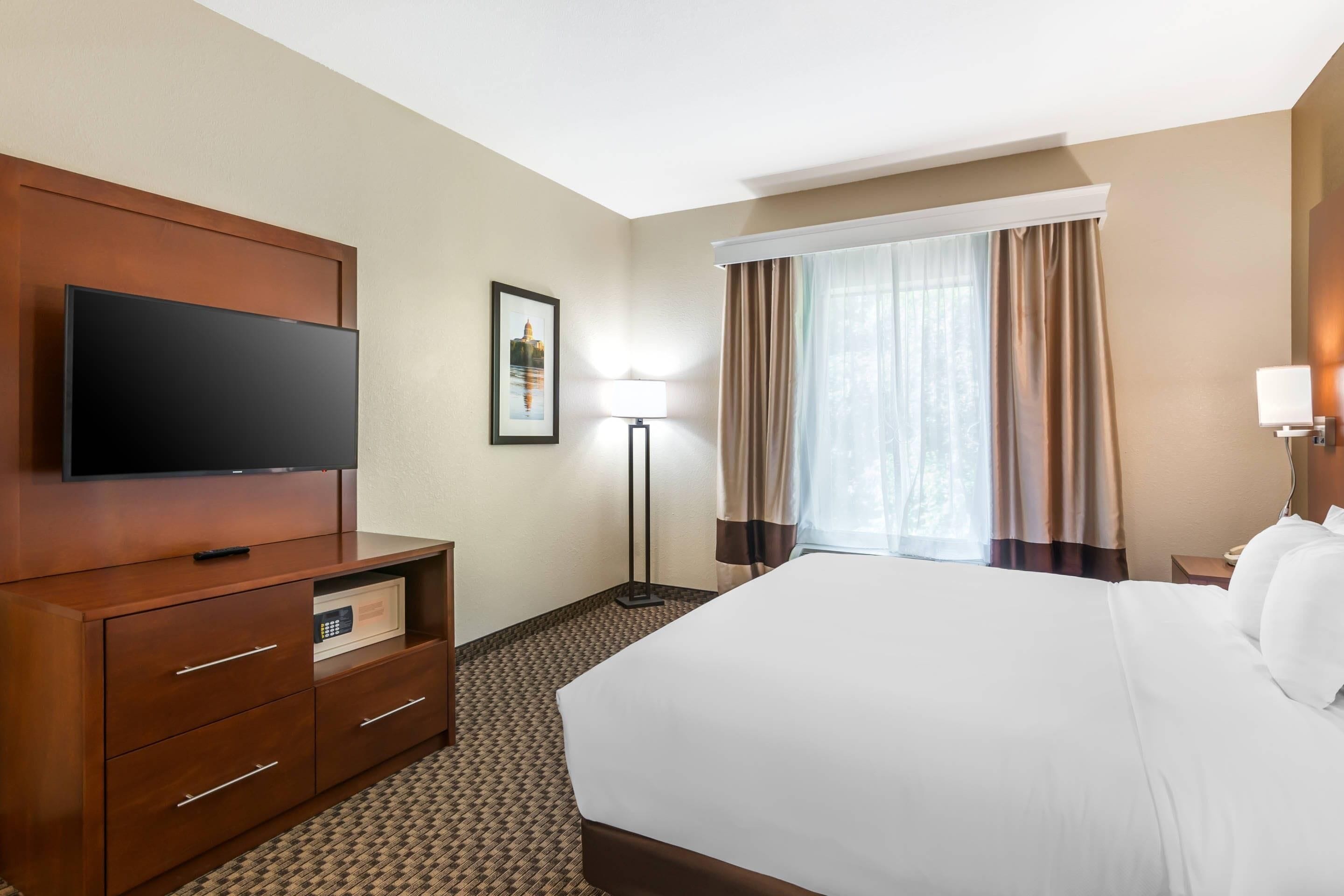 Una habitación de hotel con una cama grande, un televisor de pantalla plana sobre un aparador de madera, una lámpara de pie y una ventana con cortinas.