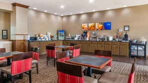 Un comedor de hotel con varias mesas y sillas, una zona de cocina con distintos electrodomésticos, y una pared decorada con cuadros.