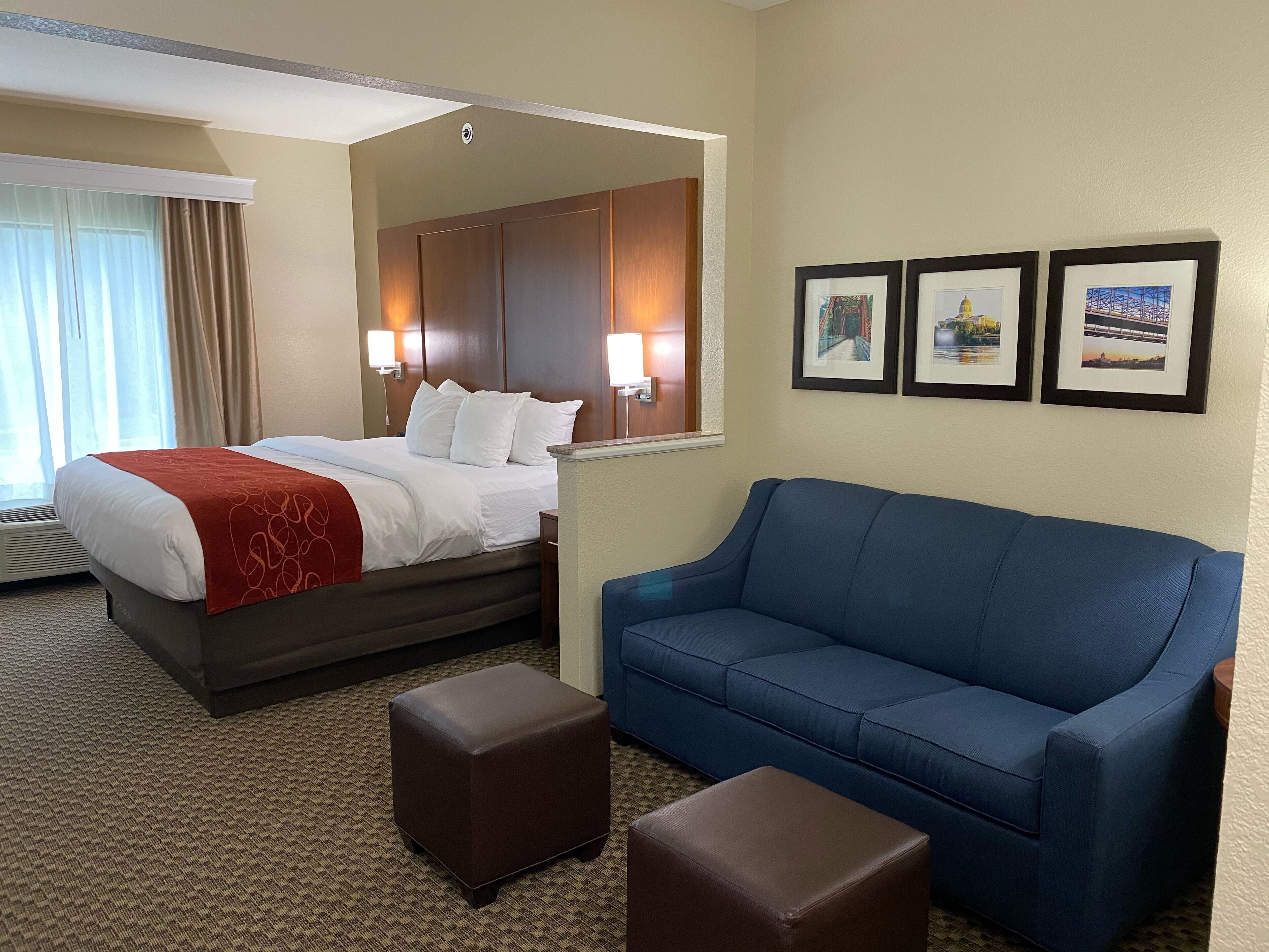 A hotel room with a bed, a blue sofa, two brown ottomans, and three framed pictures on the wall.