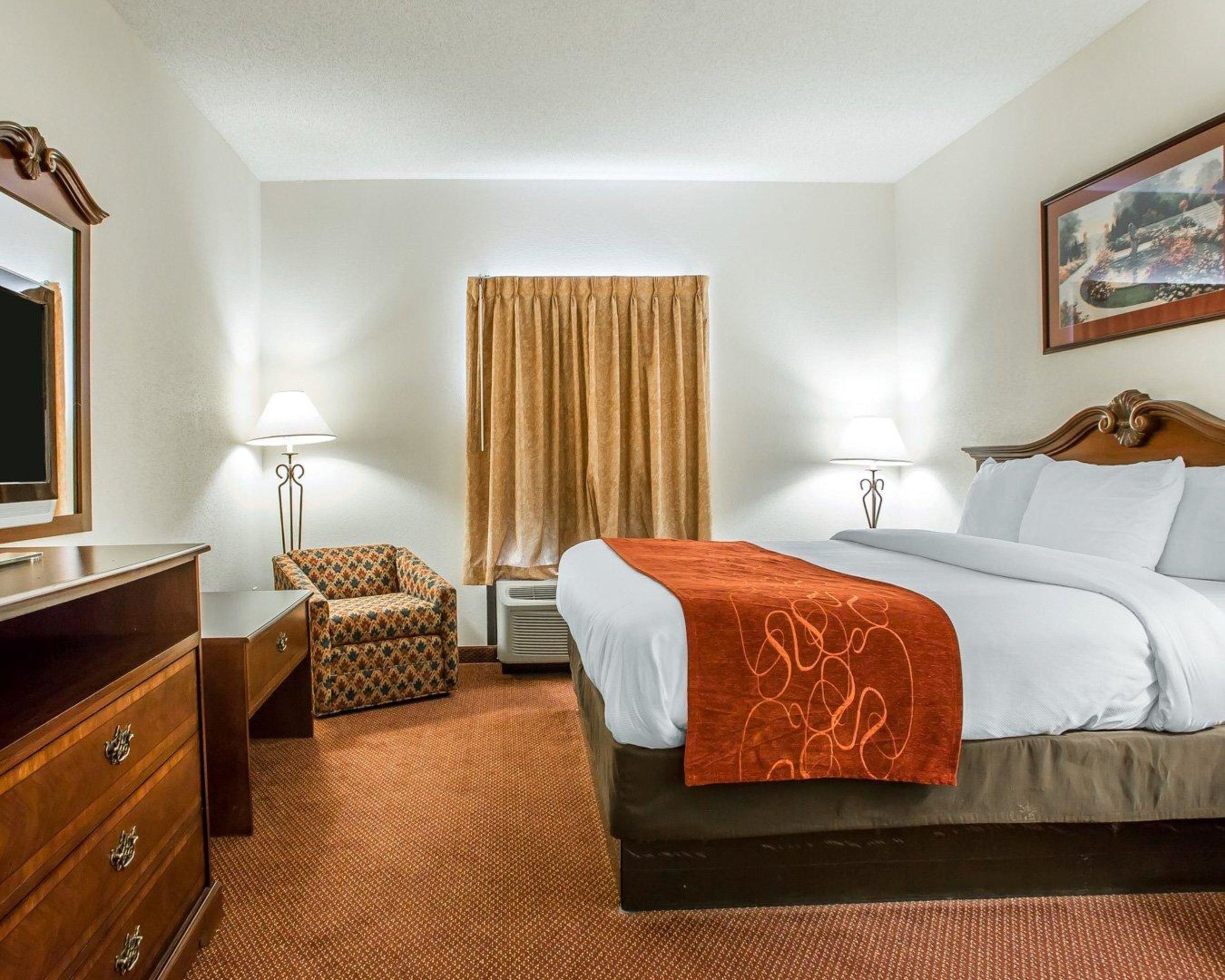 A hotel room with a bed, a dresser, a chair, a desk, a lamp, a painting, and a window with curtains.
