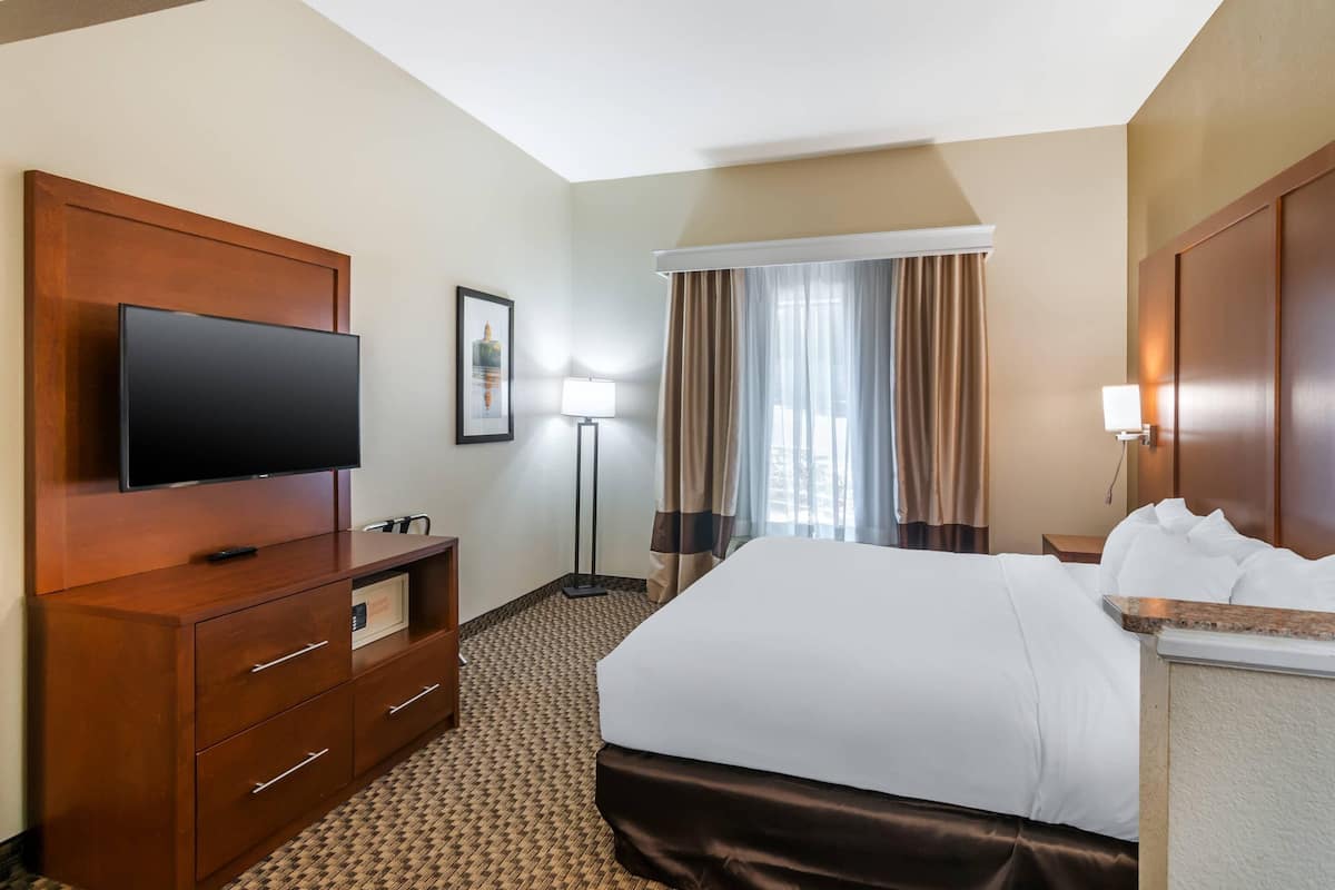 A hotel room with a large bed, a flat-screen TV mounted on a wooden cabinet, a desk with a lamp, and a window with curtains.