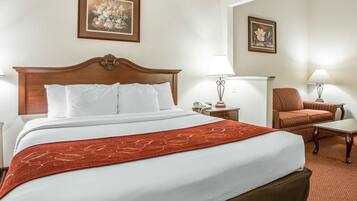 A hotel room with a large bed, a wooden headboard, two bedside tables with lamps, a chair, and two framed floral paintings on the walls.