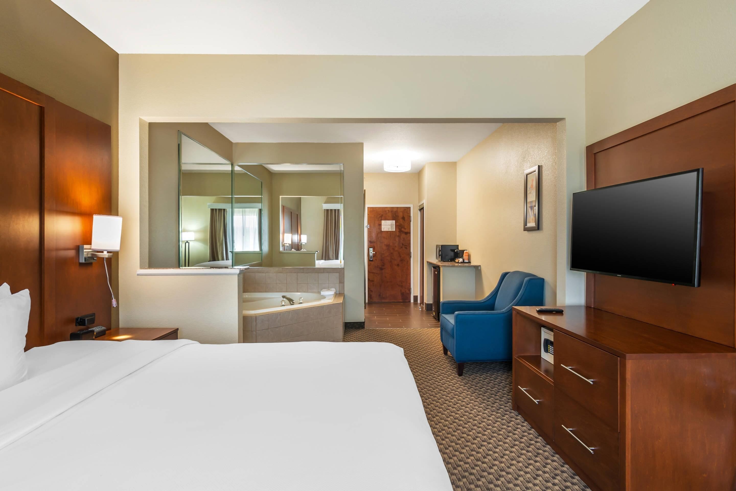 A hotel room with a large bed, a TV, a desk with a chair, a bathroom with a mirror, and a blue armchair.