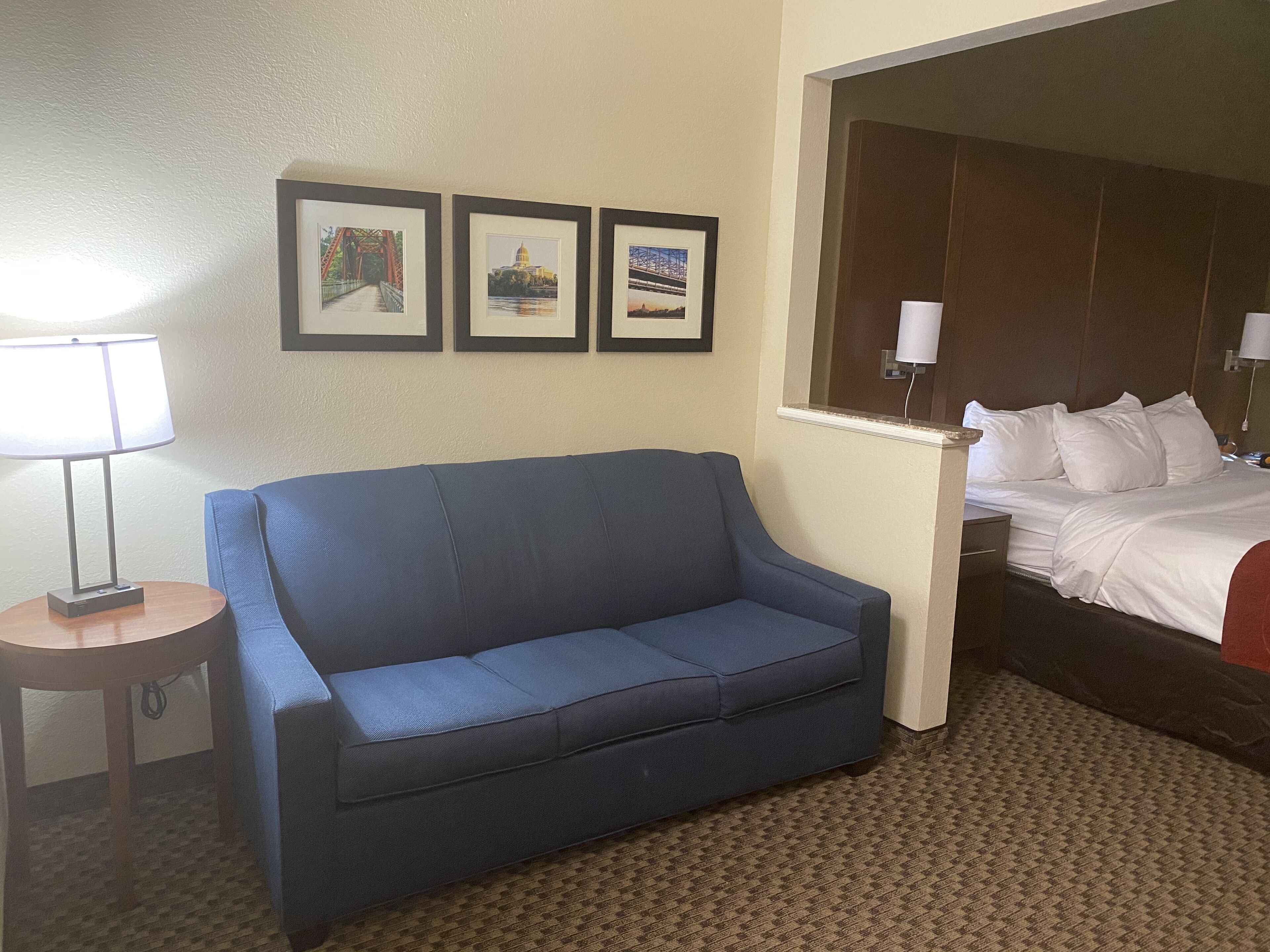 Quarto de hotel com um sofá azul, uma mesa redonda pequena com uma luminária e uma cama com roupa de cama branca. Há três quadros na parede acima do sofá.