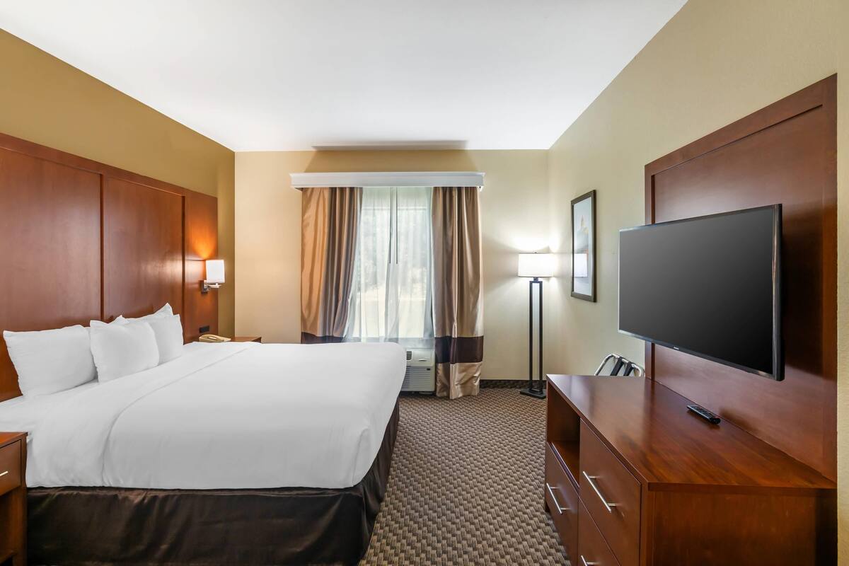A hotel room with a large bed, a flat-screen TV, a desk with a lamp, and a window with curtains.