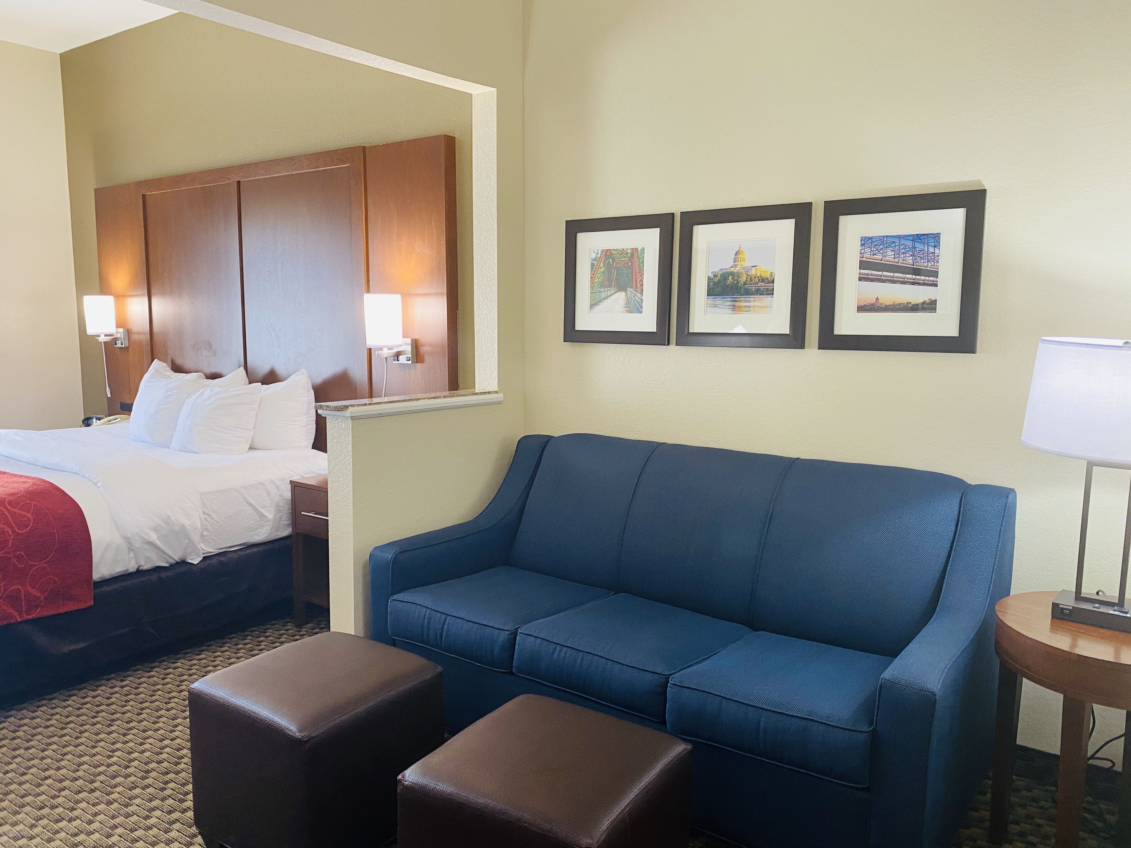 Une chambre d’hôtel avec un lit, un canapé bleu, deux poufs marron, une petite table avec une lampe et trois tableaux encadrés accrochés au mur.