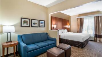 A hotel room with a blue sofa, a small round table, a bed with a white comforter, and a window with curtains.