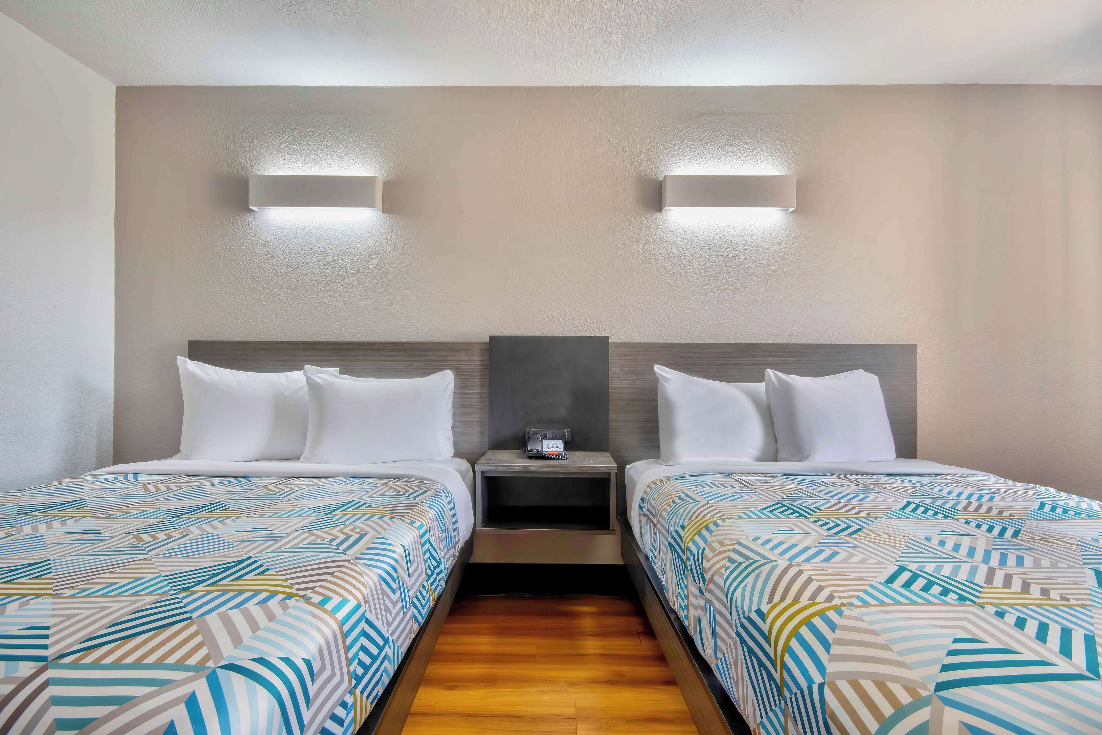 A hotel room with two beds, a nightstand between them, and wall-mounted lighting.