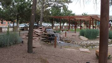 Un parque con una pérgola de madera, una cascada pequeña y un estacionamiento con coches.