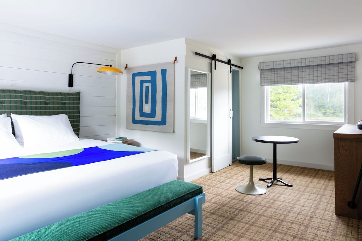 Habitación de hotel con cama, banca, mesita, lámpara y ventana con vista a follaje.
