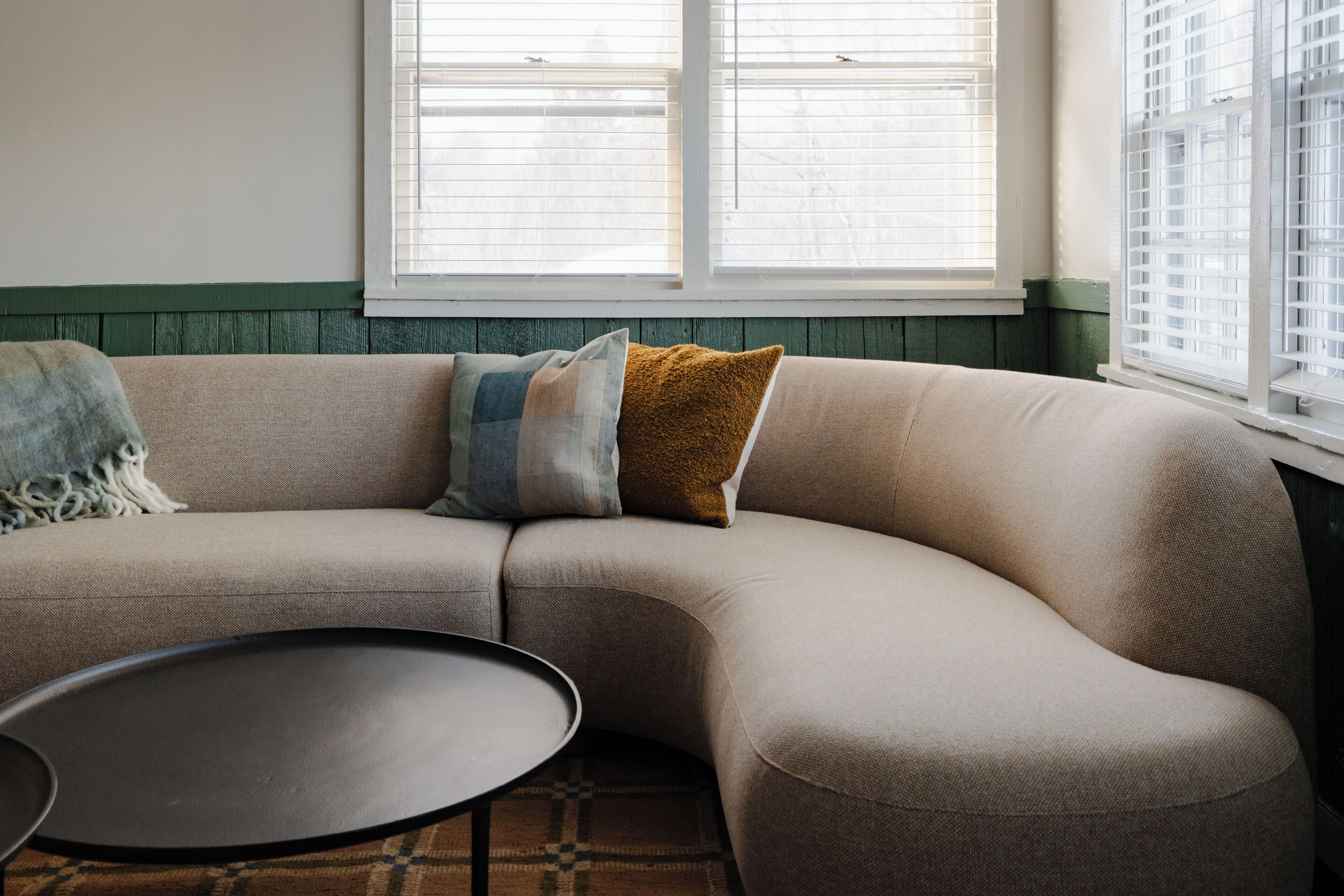 A beige sectional sofa with a patterned throw, a small round table, and a window with blinds.