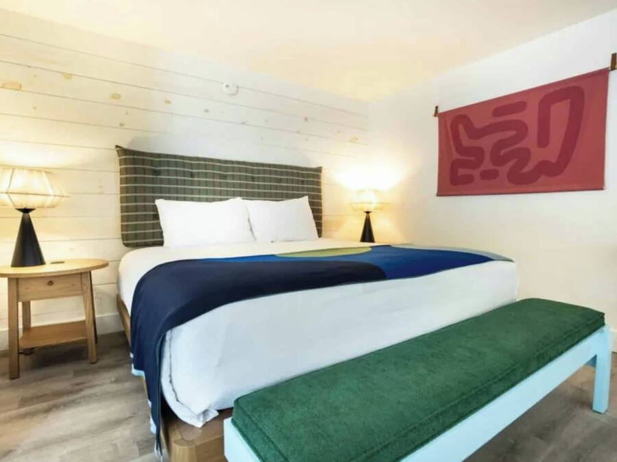 Un dormitorio con cama, mesitas de noche, un banco y una obra de arte colgada en la pared.