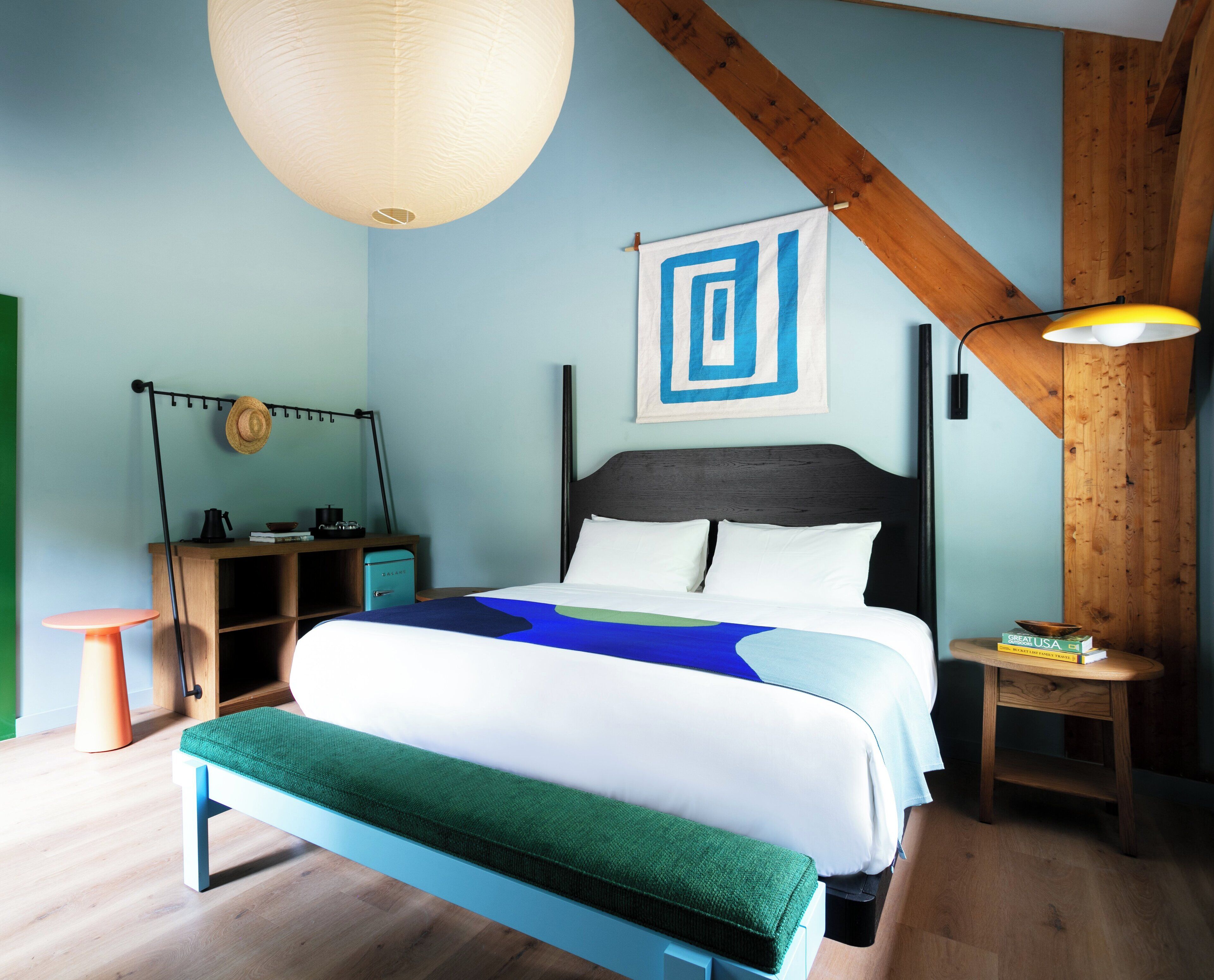 A bedroom with a bed, a bedside table, a lamp, a hat on the wall, and a painting.
