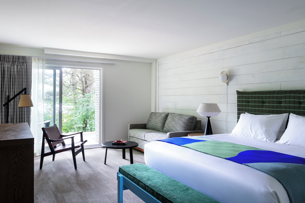 A hotel room with a bed, a sofa, a chair, a small table, and a lamp.