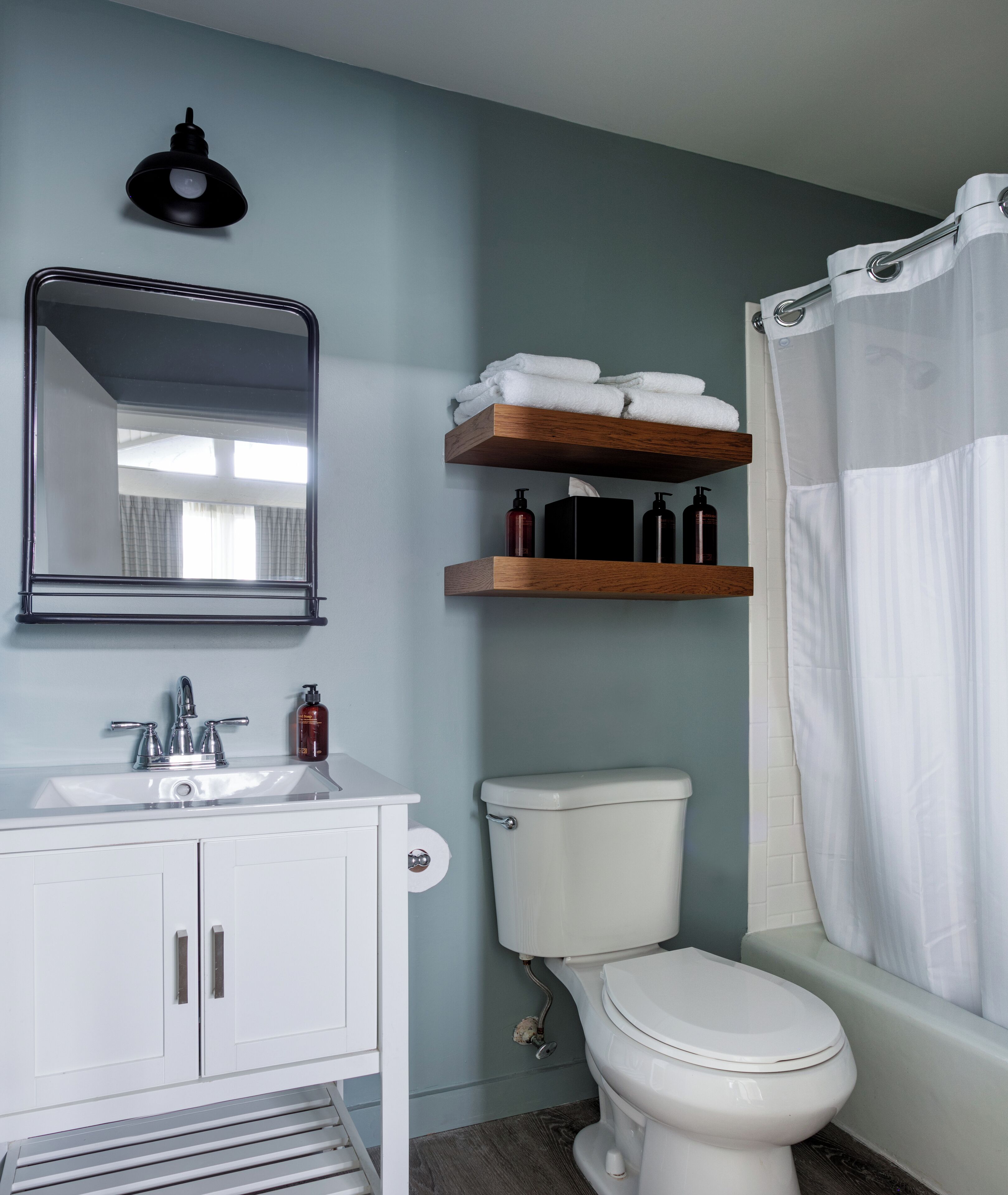 A bathroom with a white sink, mirror, and toilet. There are wooden shelves above the toilet with toiletries and a white towel.