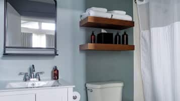 Ein Badezimmer mit Waschbecken, Spiegel und Toilette. Über der Toilette befinden sich Holzregale mit Toilettenartikeln und einem weißen Handtuch.