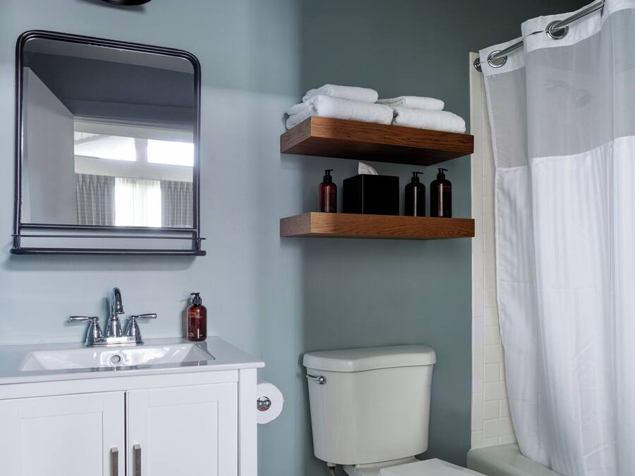 Ein Badezimmer mit Waschbecken, Spiegel und Toilette. Über der Toilette befinden sich Holzregale mit Toilettenartikeln und einem weißen Handtuch.