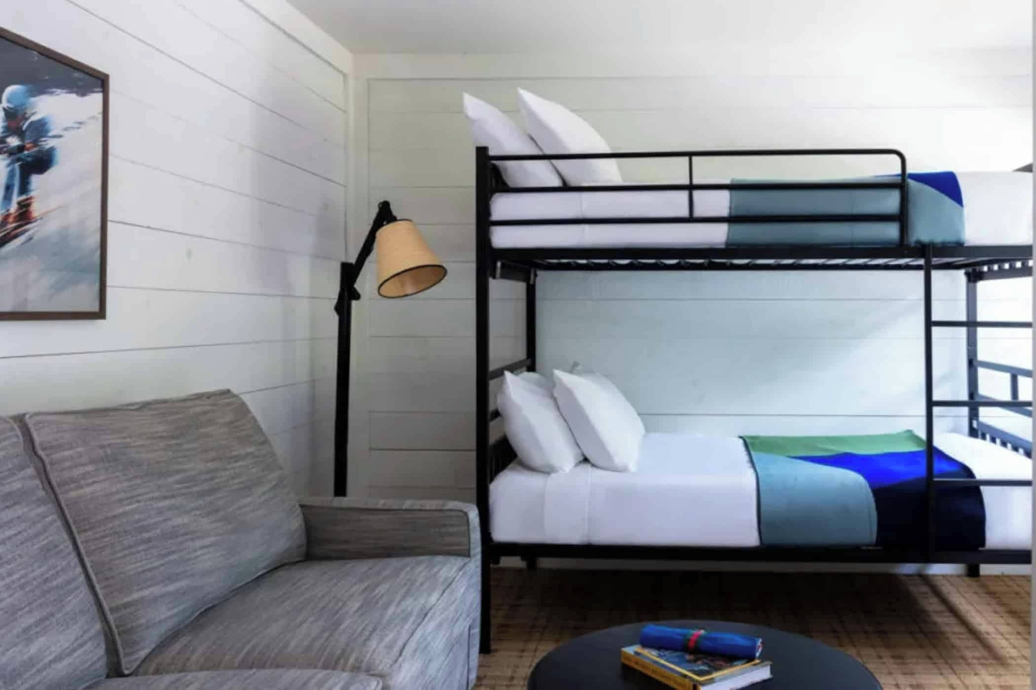 A bunk bed with white bedding, a grey sofa, a floor lamp, and a small table with books.