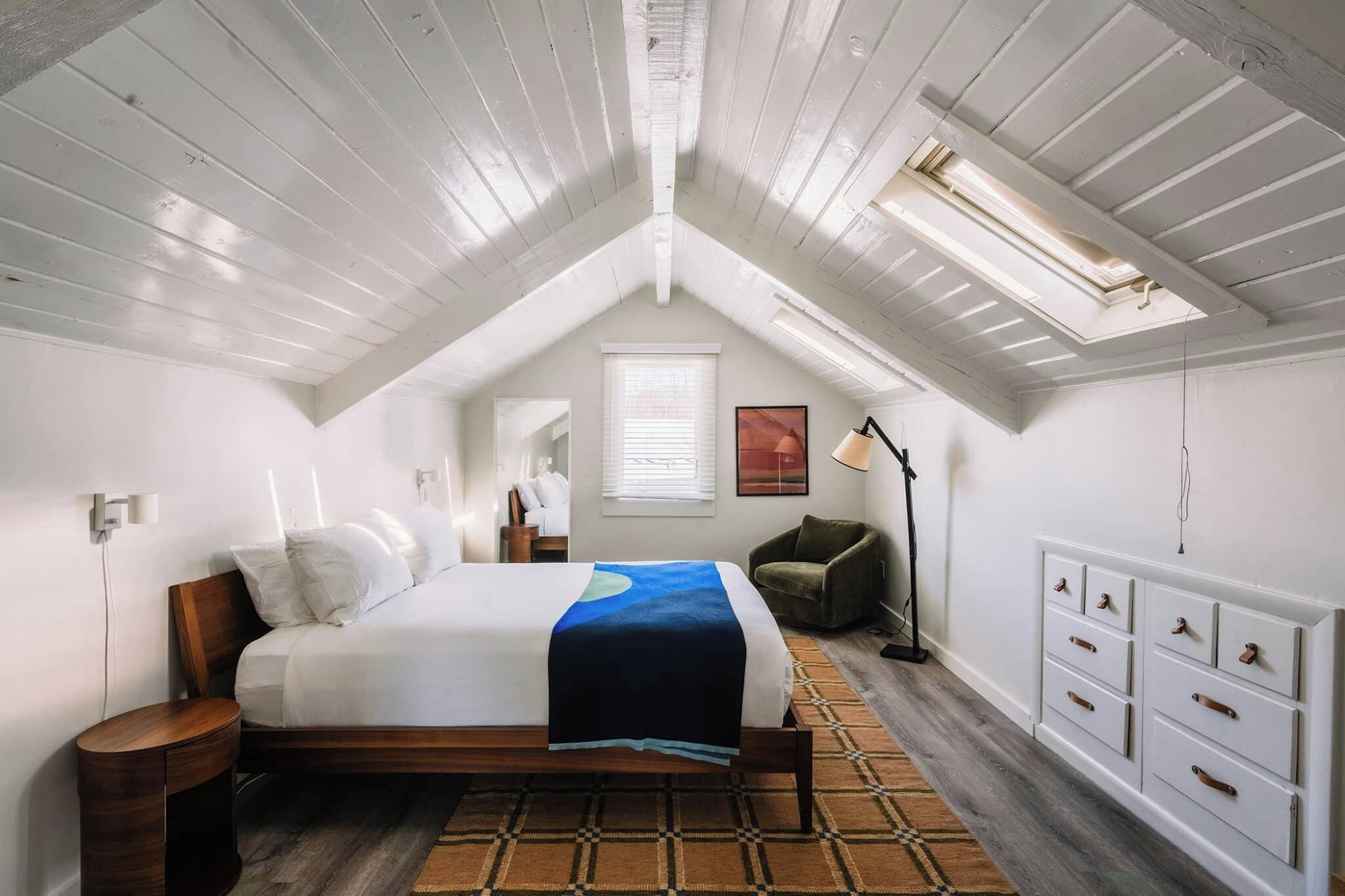 A bedroom with a sloped ceiling, a bed with a blue blanket, a chair, a floor lamp, and a dresser.