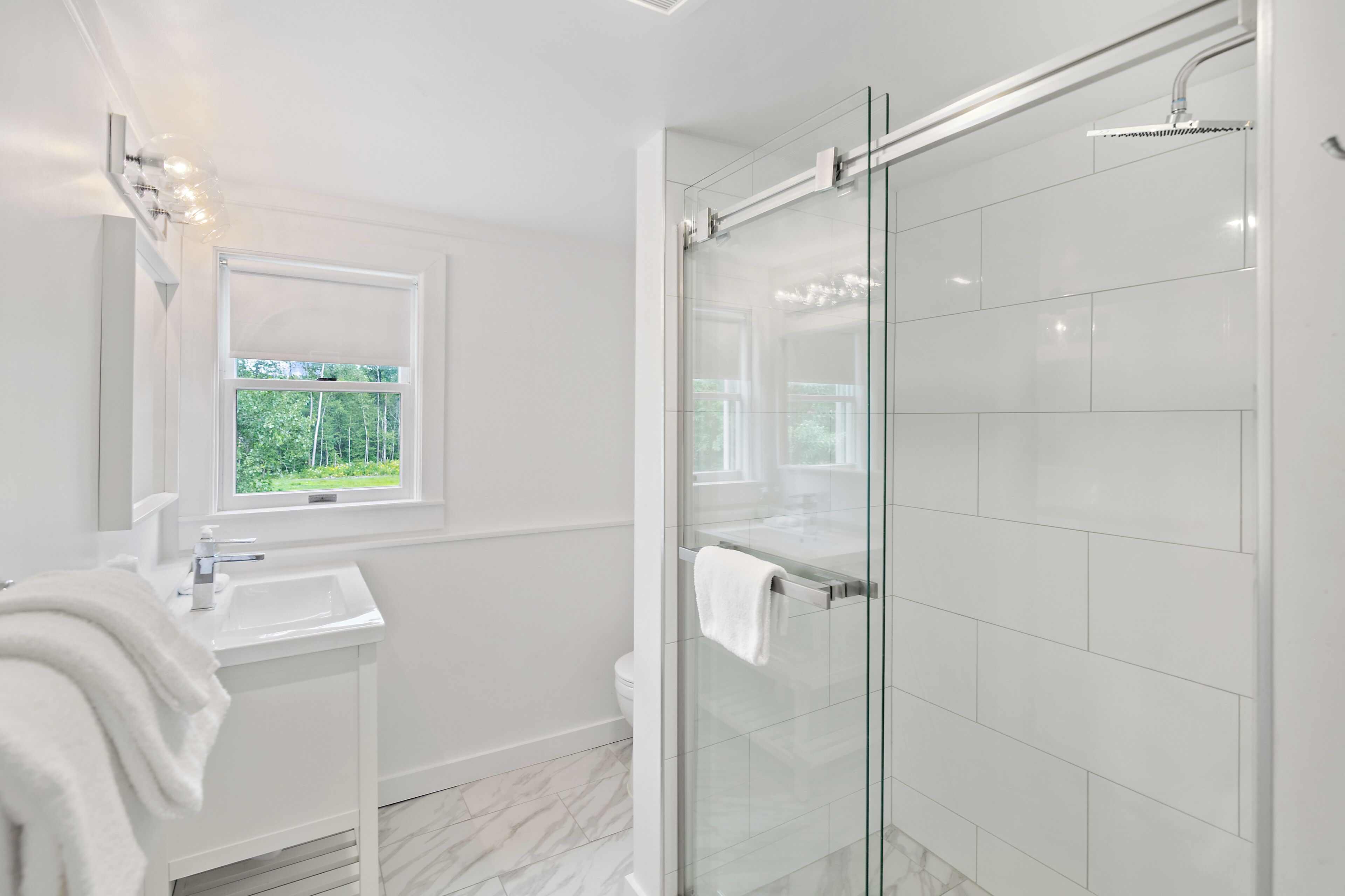 Un baño moderno con ducha de vidrio, lavamanos blanco y ventana con vistas a la vegetación.