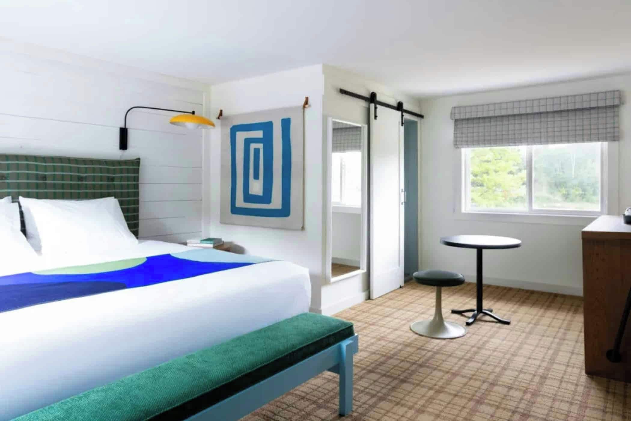 A hotel room with a bed, bedside table, lamp, artwork, window, and a small round table.