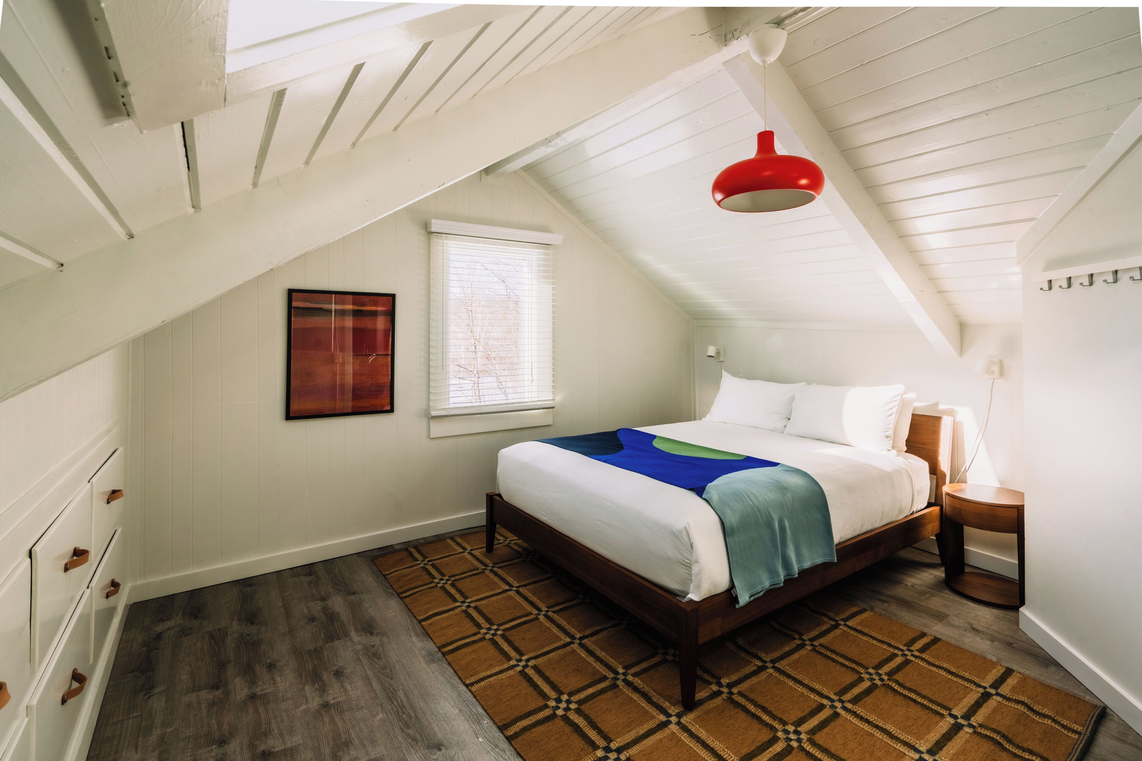 Un dormitorio con cama, ventana, lámpara colgante roja y alfombra estampada.
