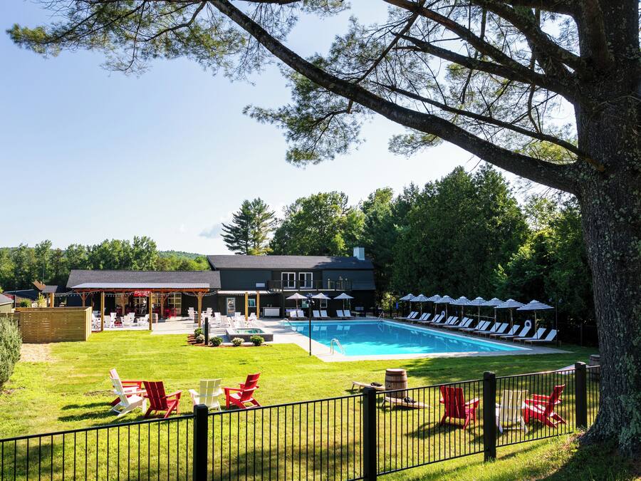 A pool area with lounge chairs and umbrellas, a building with a covered patio, and a fenced lawn.