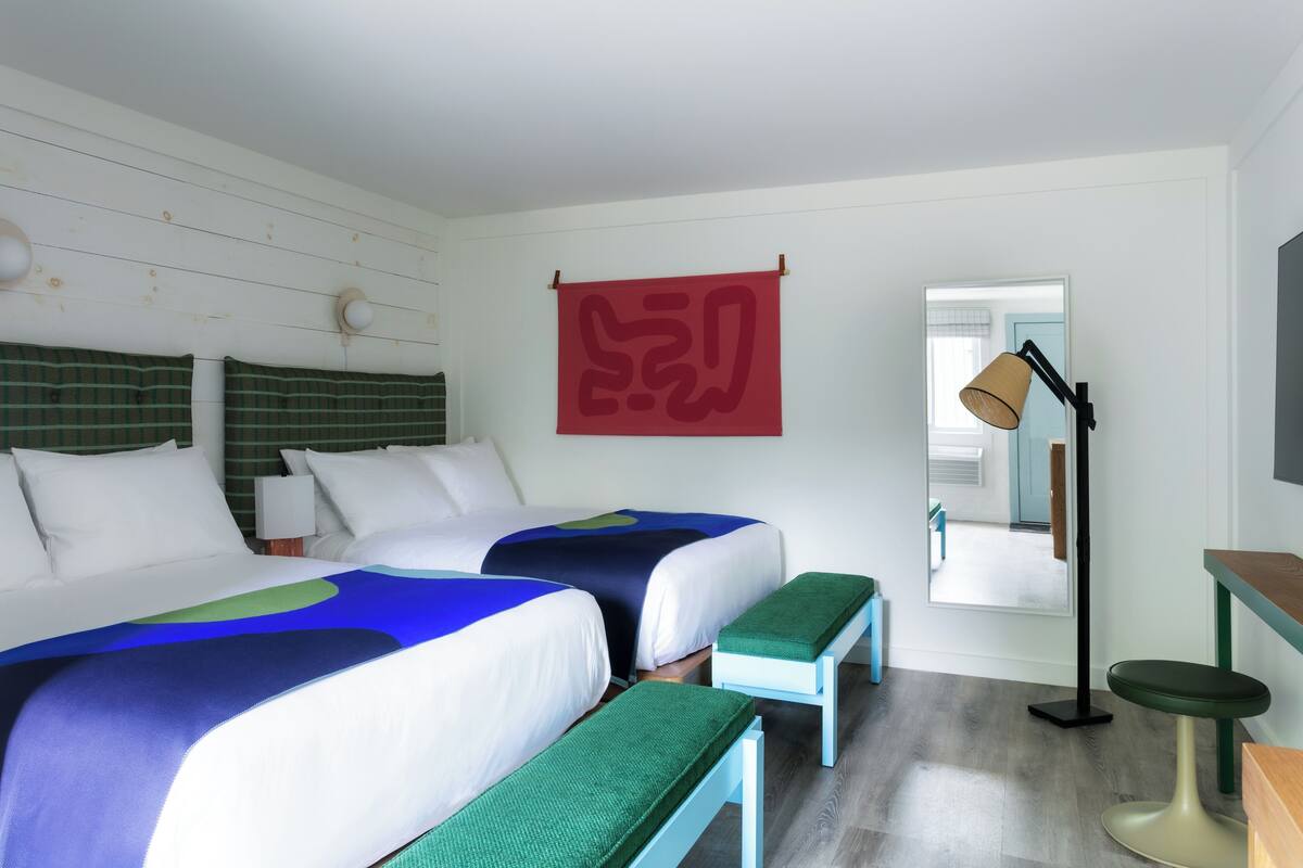 Una habitación de hotel con dos camas, un escritorio, una silla, una lámpara y un espejo grande.