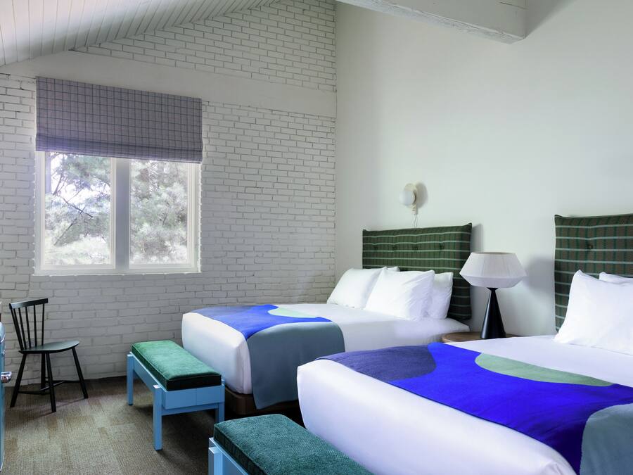 A hotel room with two beds, a desk, a chair, and a window with a view of trees.