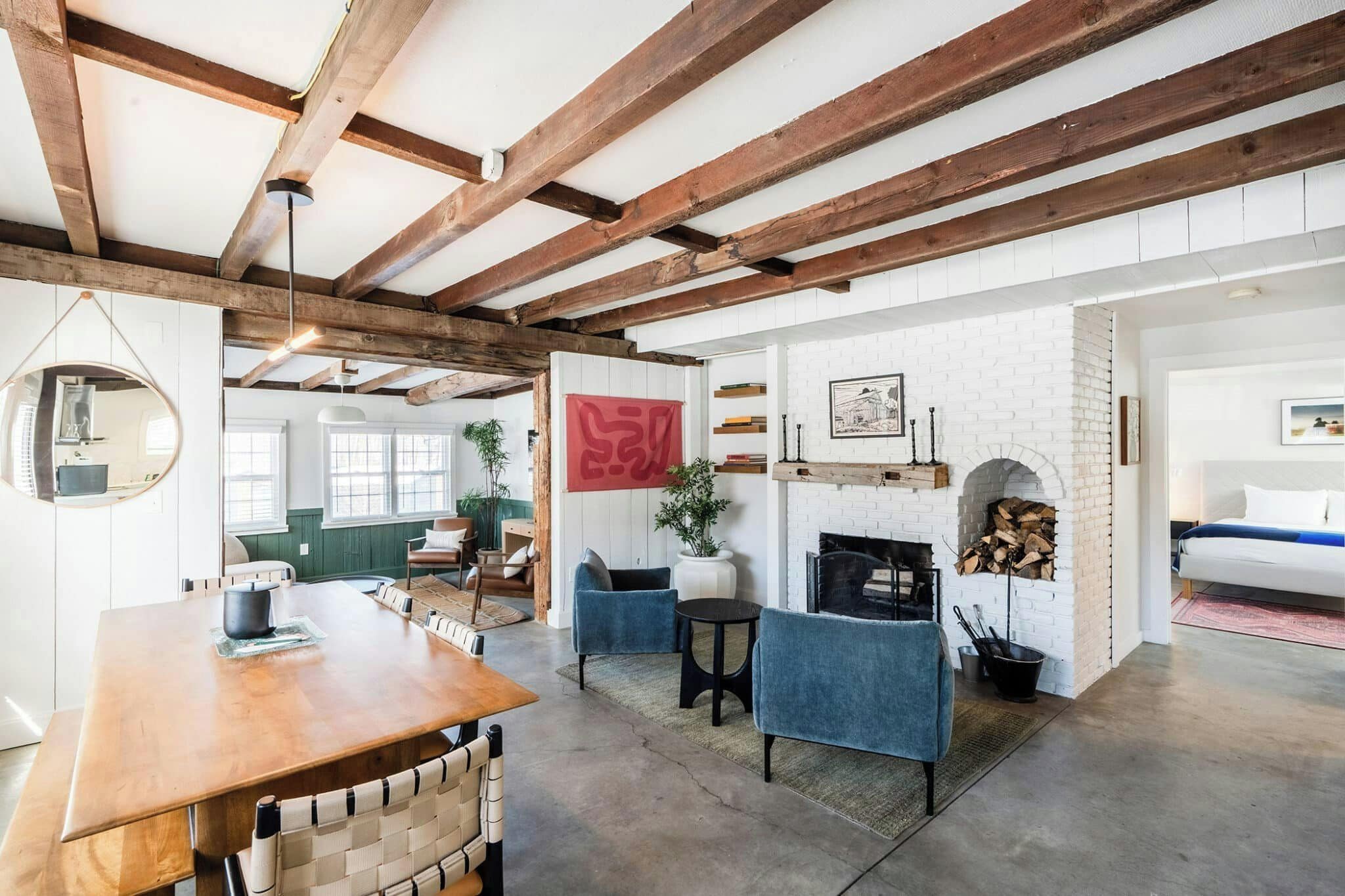 A spacious room with exposed wooden beams, a fireplace, a round mirror, and a dining area.