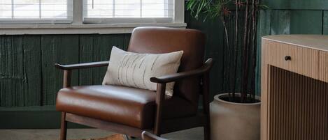 Una habitación con una silla de cuero marrón, una mesita de madera, una pared verde, una ventana con persianas y una planta en maceta.