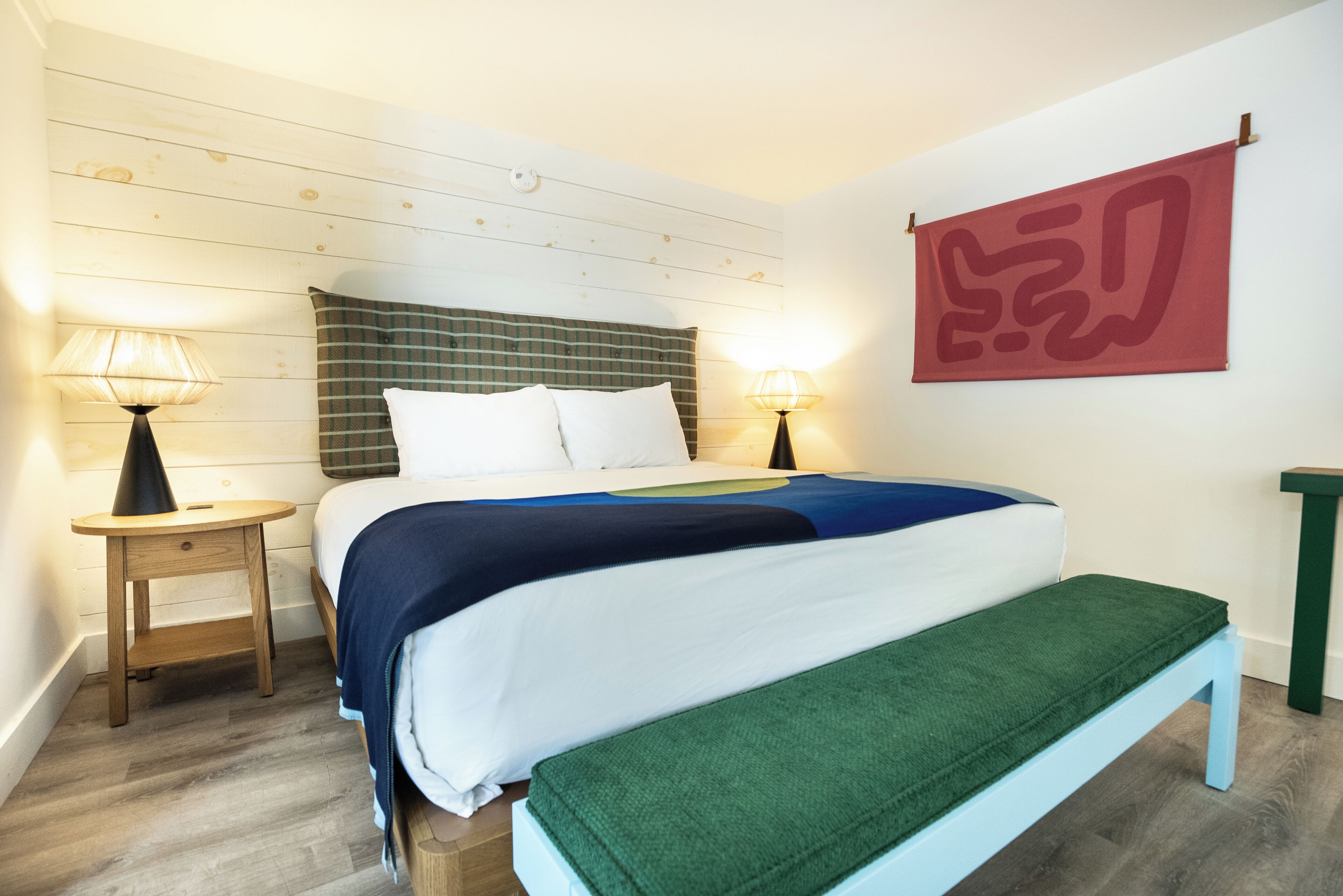 A neatly arranged bedroom with a bed, two bedside lamps, a bench, and a wall-mounted artwork.