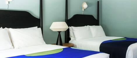 Habitación de hotel con dos camas, cada una con cabecero blanco y colcha abstracta azul y verde. Se observa una mesita de noche con lámpara y un aplique de pared.