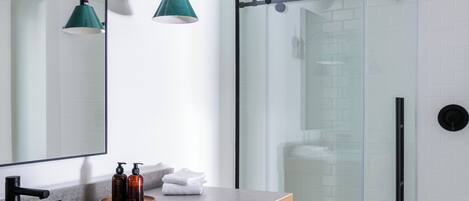 Ein modernes Badezimmer mit einem Holzkosmetikschrank, einem großen Spiegel, zwei Wandleuchten, einer Duschkabine aus Glas und einem gefliesten Boden.