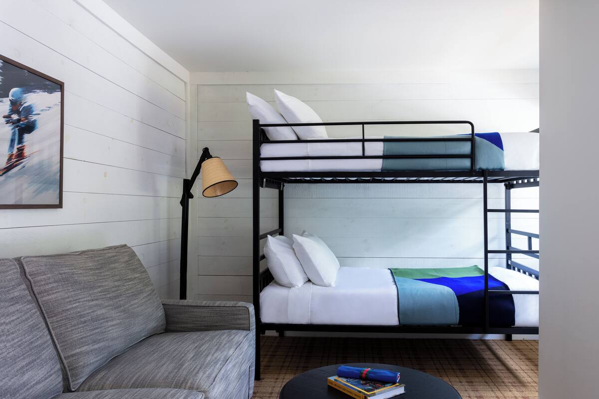 Una cama litera con ropa blanca, un sofá gris y una lámpara de pie.