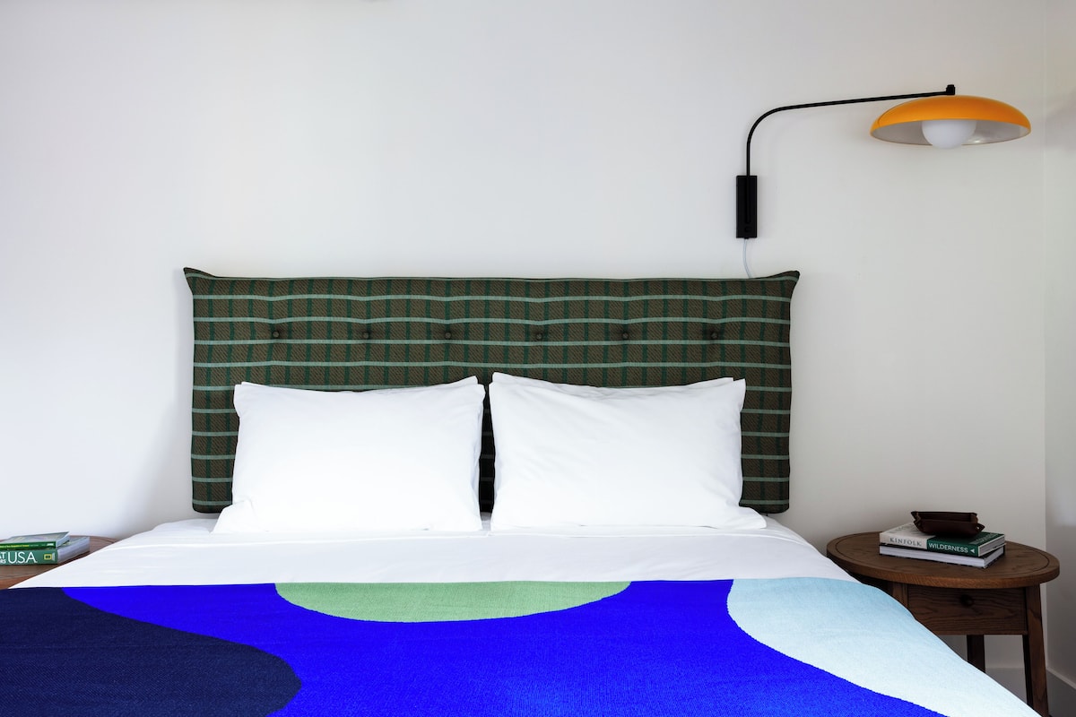 Una cama bien hecha con una cabecera de cuadros verdes, almohadas blancas y una manta azul y verde. Hay una mesita de noche de madera con una lámpara y algunos libros.