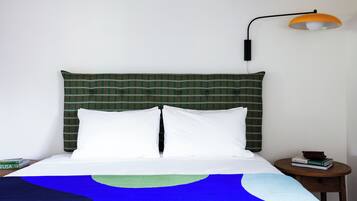 Una cama bien hecha con una cabecera de cuadros verdes, almohadas blancas y una manta azul y verde. Hay una mesita de noche de madera con una lámpara y algunos libros.