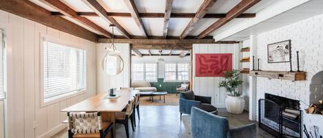 Een ruime kamer met een houten plafond, een zithoek met stoelen, een bank, een open haard en een groot raam.