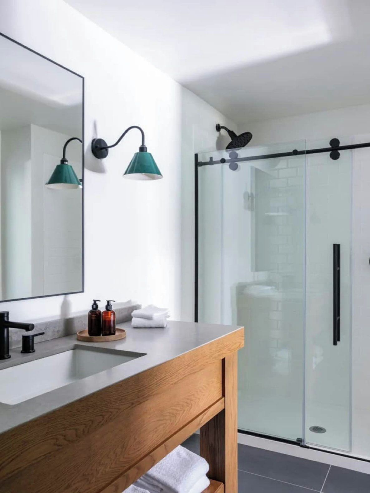 Een moderne badkamer met een houten wastafelmeubel, een grote spiegel, twee wandlampen, een glazen douchecabine en een handdoekrek.
