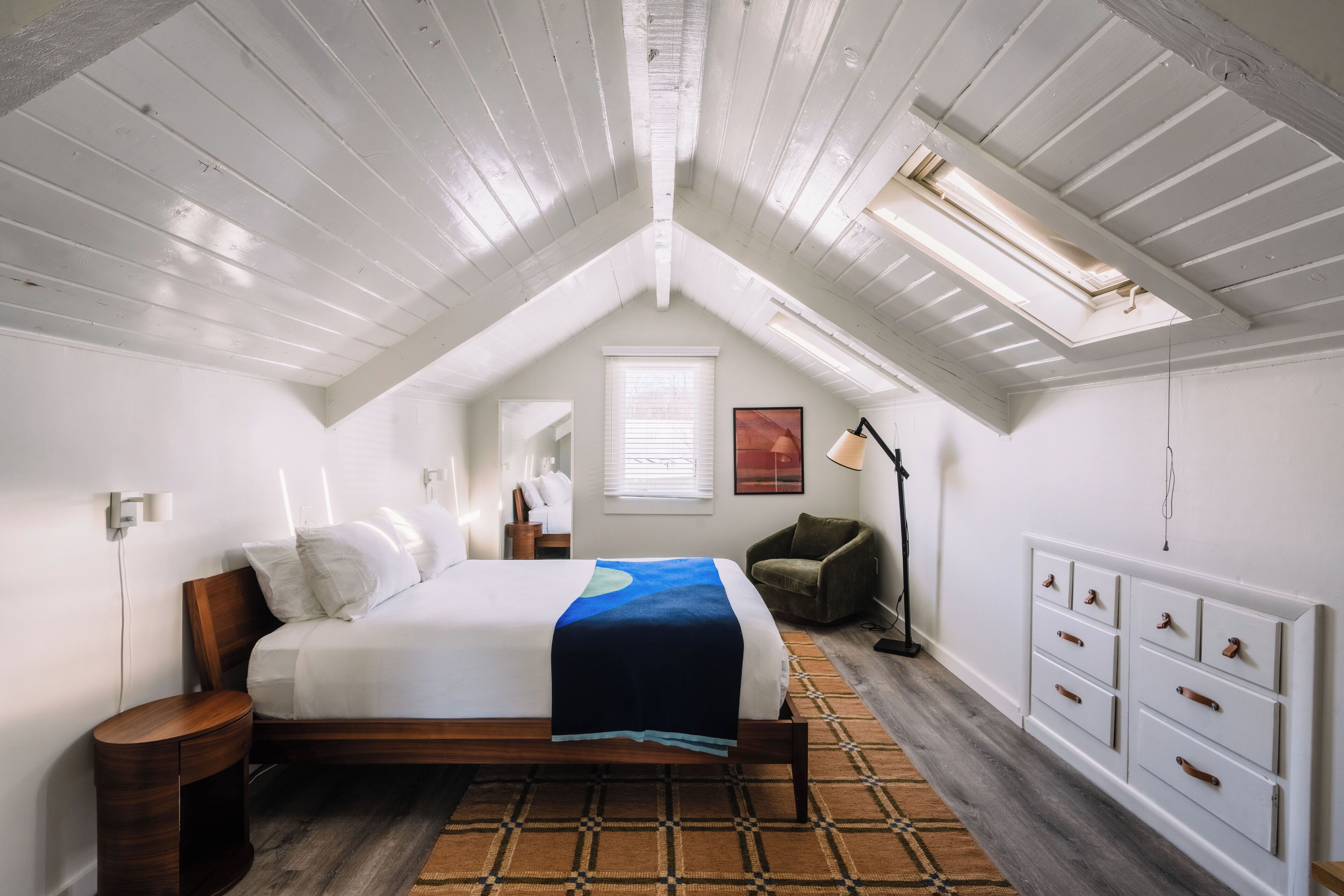 A bedroom with a sloped ceiling, a bed, a nightstand, a chair, a dresser, and a window.