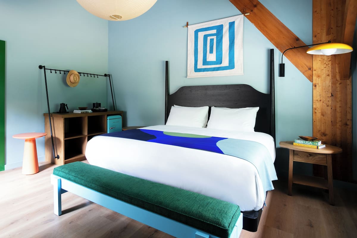 Un dormitorio con cama, mesita de noche, lámpara, un sombrero en la pared y un marco de fotos.