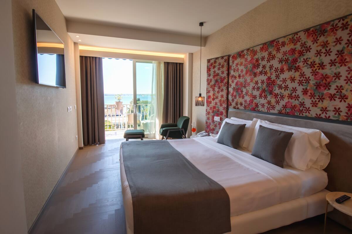 A hotel room with a large bed, a flat-screen TV mounted on the wall, a balcony with a view of the sea, and a floral-patterned headboard.