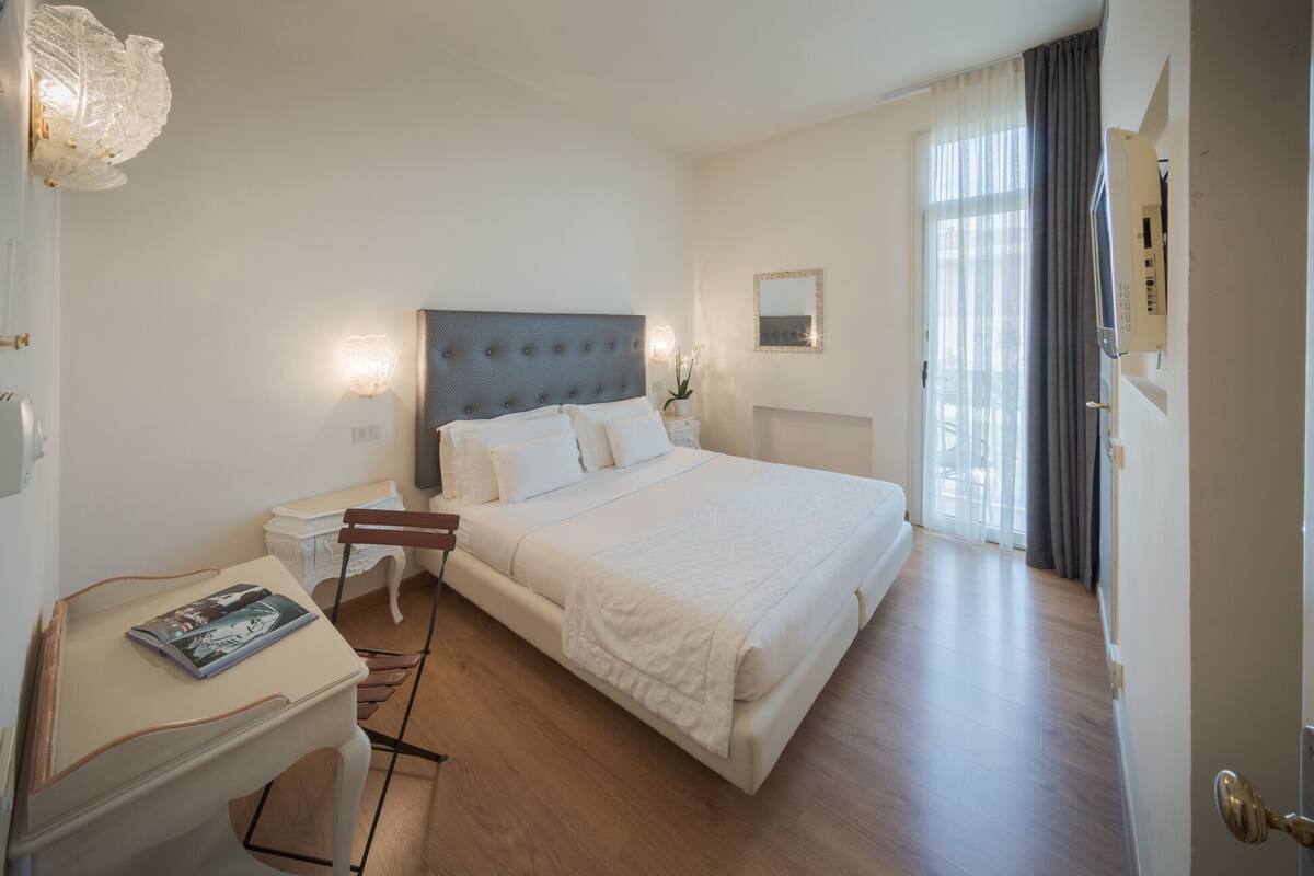 A hotel room with a large bed, a bedside table with a lamp, a chair, and a window with curtains.