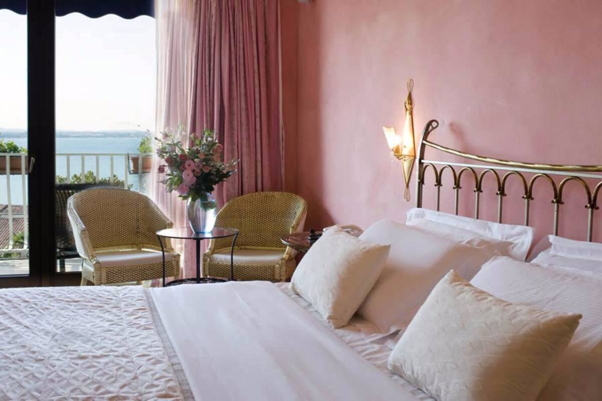 A hotel room with a bed, two wicker chairs, a small table with a vase, and a view of the sea.