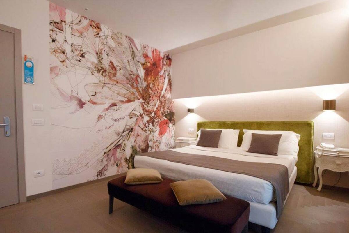 A modern hotel room with a large floral artwork on the wall, a neatly made bed with two pillows, a bench, and a nightstand with a lamp.