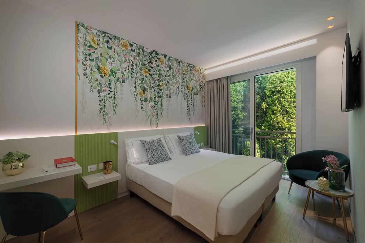 A modern hotel room with a large bed, a green chair, a small table with a vase, and a view of greenery through the window.