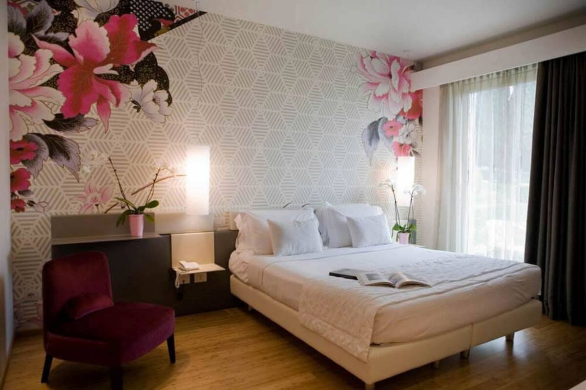 A hotel room with a large bed, a red chair, a nightstand, and a wall with a floral pattern.
