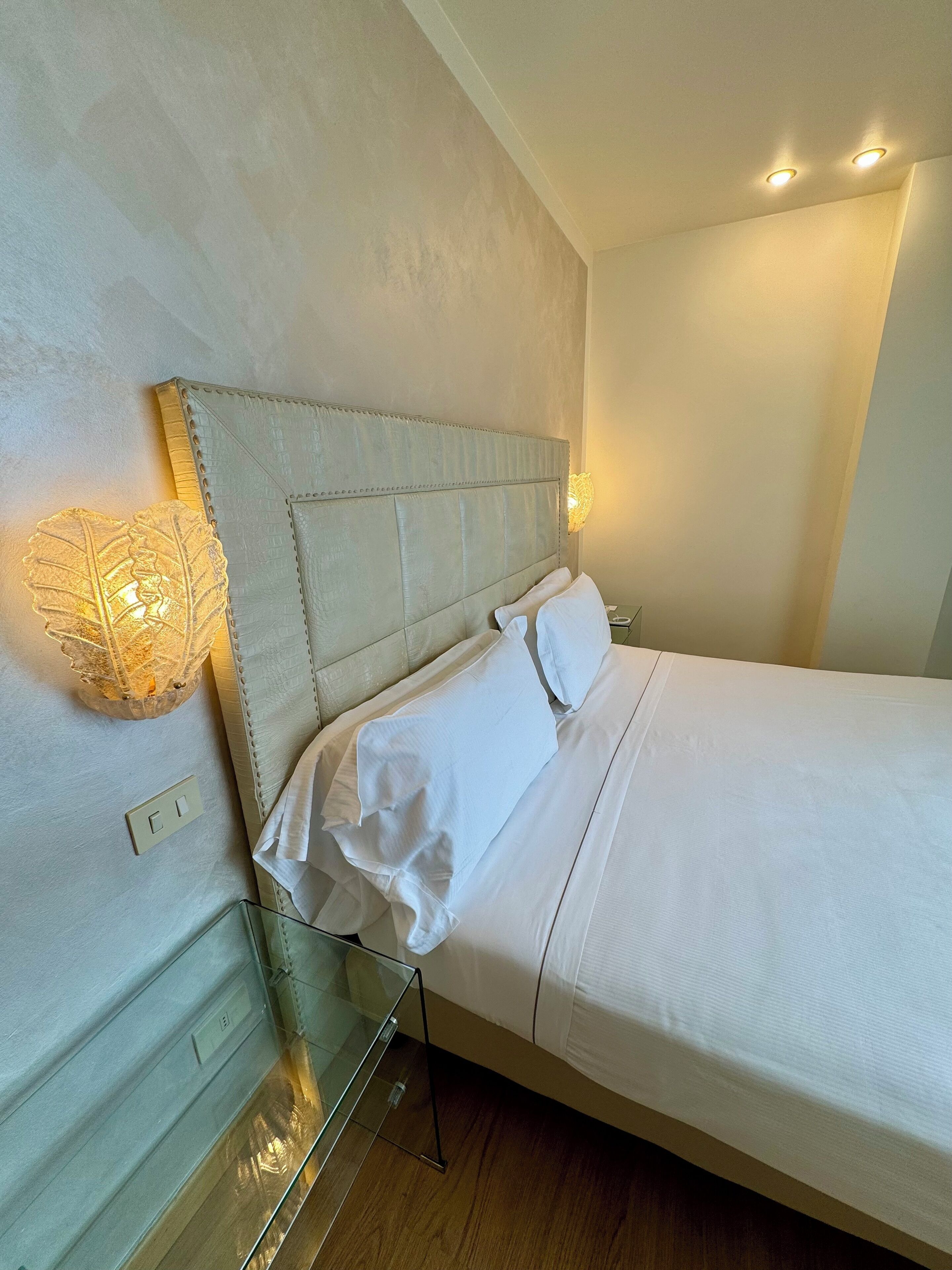 Uma cama bem arrumada com lençóis brancos, cabeceira acolchoada e luminária de cabeceira.