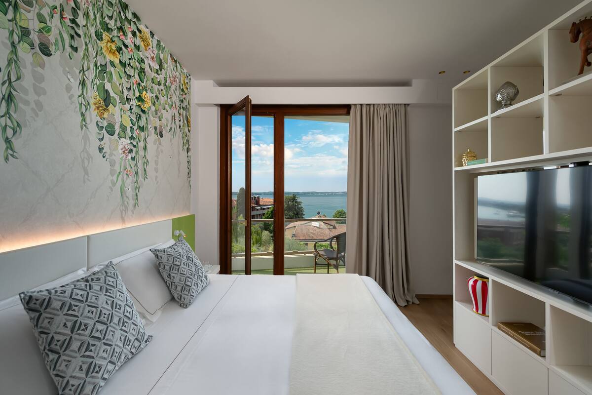 A bedroom with a large bed, a TV, and a balcony with a view of the sea.